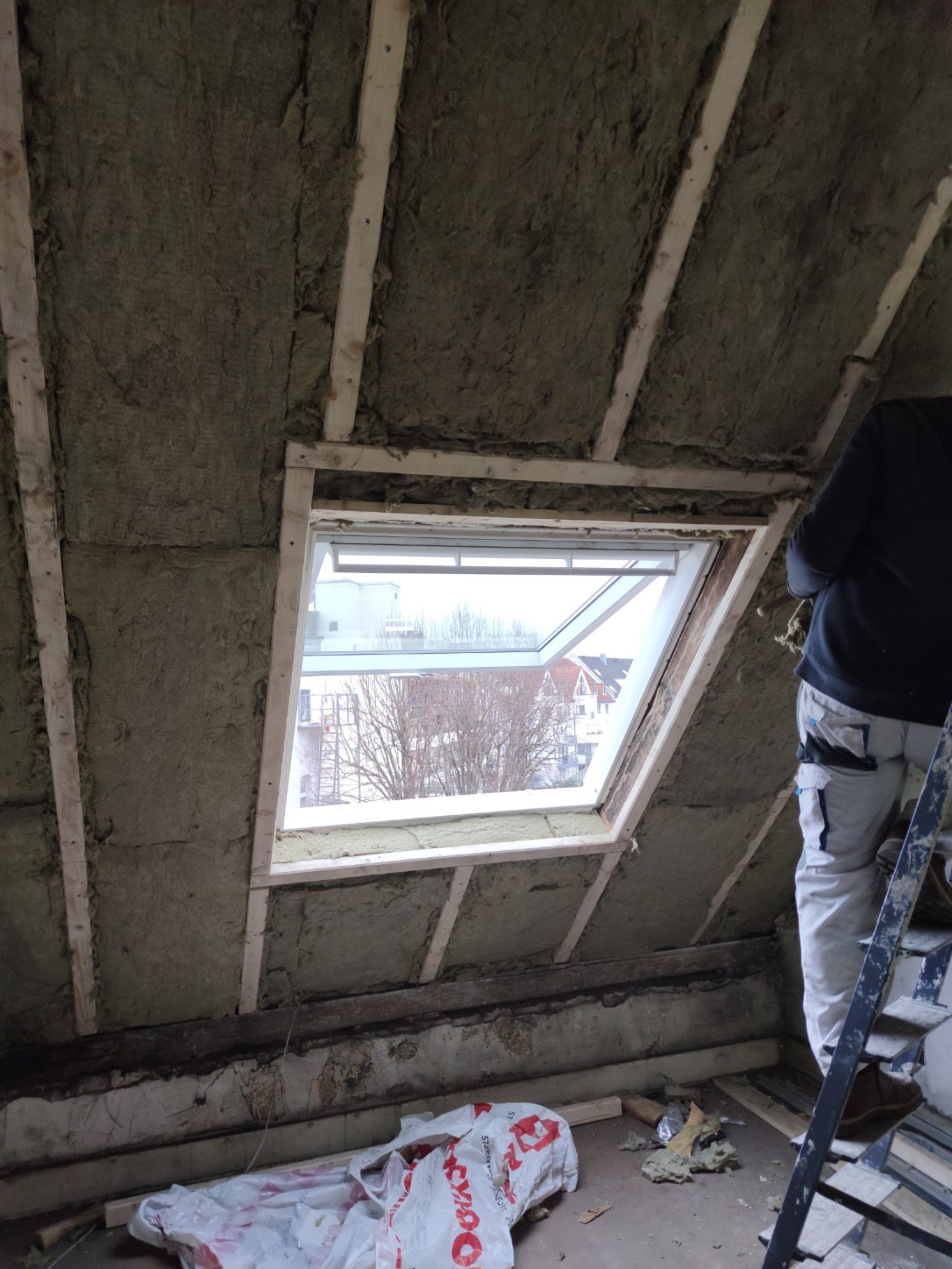 Ein unvollendetes Zimmer mit einer Dachfensteröffnung und einer Person auf einer Leiter.